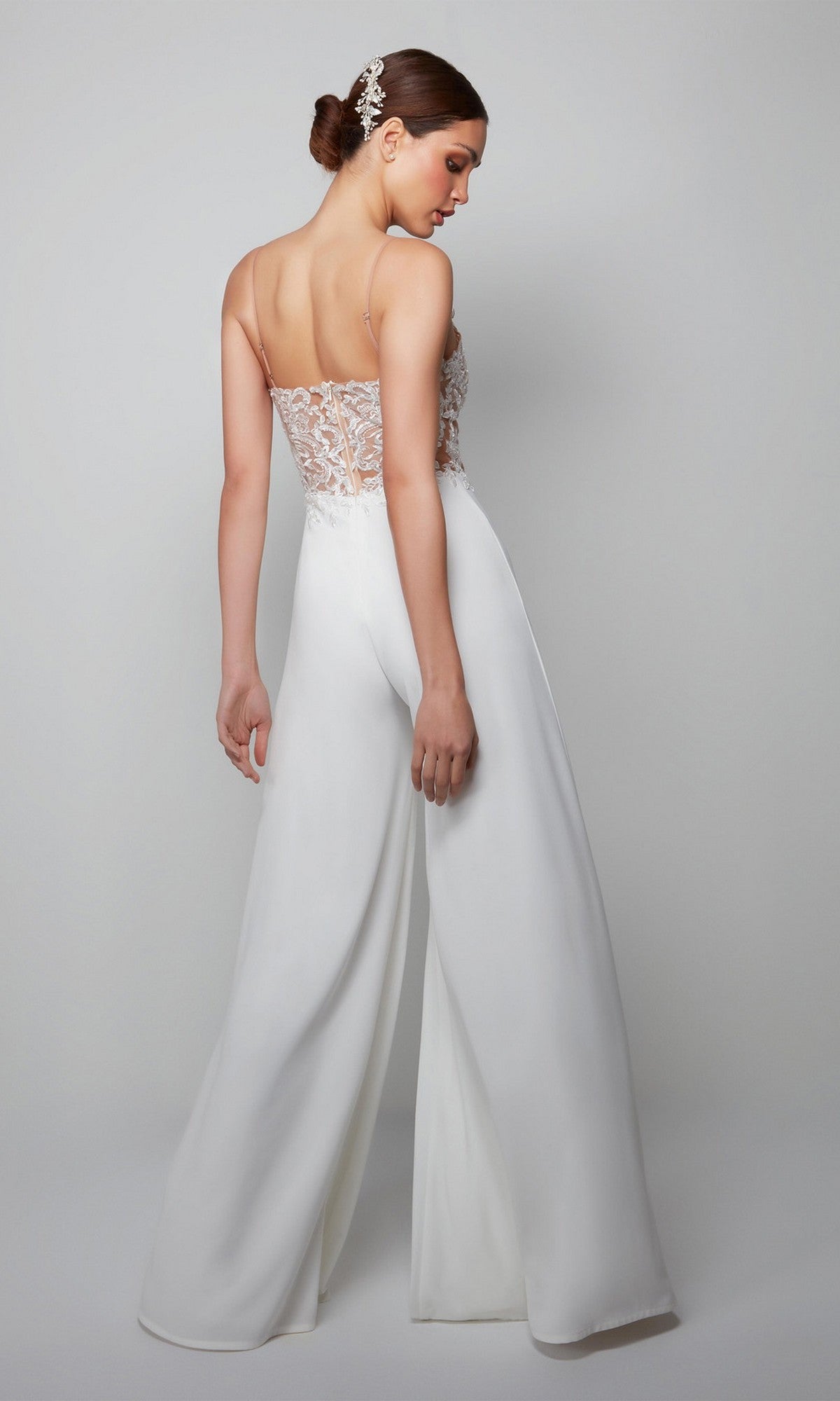 A woman poses gracefully with her back turned, wearing the Alyce 70017 long white jumpsuit featuring a sheer lace bodice and wide-leg pants.